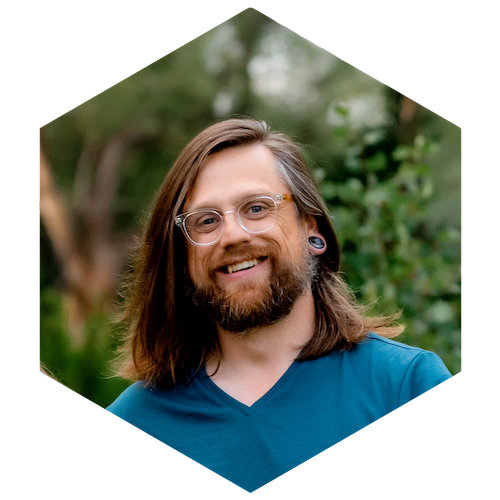 A smiling person with shoulder length brown hair, greying temples, and a mixed red-brown-grey beard. They are wearing a teal v-neck t-shirt, wood and clear framed glasses, and large guage black stone ear jewlery. The background is an out-of-focus nature setting.